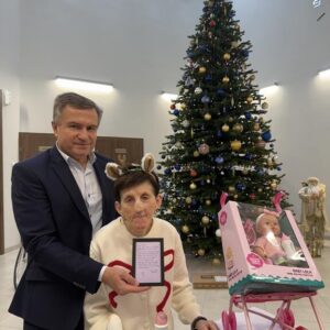 Mężczyzna w garniturze stoi obok starszej kobiety w swetrze z reniferem, trzymając kartkę z życzeniami. Na stole znajdują się kosmetyki, a obok nich wózek dla lalki z dzieckiem. W tle widać udekorowaną choinkę.