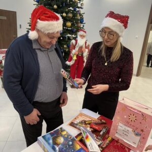 Mężczyzna i kobieta w czapkach świątecznych rozmawiają o grach planszowych na tle choinki i dekoracji bożonarodzeniowych. Na stole widoczne są różnorodne gry i upominki, a w tle stoi Mikołaj.