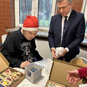 Mężczyzna w garniturze wręcza prezent młodemu mężczyźnie w czapce Mikołaja, którzy siedzą przy stole w jasnym pomieszczeniu. Na stole znajdują się kolorowe zestawy edukacyjne i otwarte pudełka z różnymi materiałami. W tle widać okna z widokiem na las.