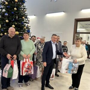 Grupa osób stojąca przed choinką świąteczną, trzymająca kolorowe torby z prezentami. W tle widoczna dekoracja wnętrza oraz zegar na ścianie. Uczestnicy wydarzenia uśmiechają się, celebrując atmosferę radości i wspólnoty w okresie świątecznym.