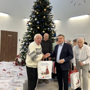 Uczestnicy wydarzenia wręczają prezenty z okazji świąt Bożego Narodzenia, stojąc przed dużą choinką w tle. Widać torby z darami i uśmiechnięte twarze, co podkreśla radosną atmosferę świąteczną.