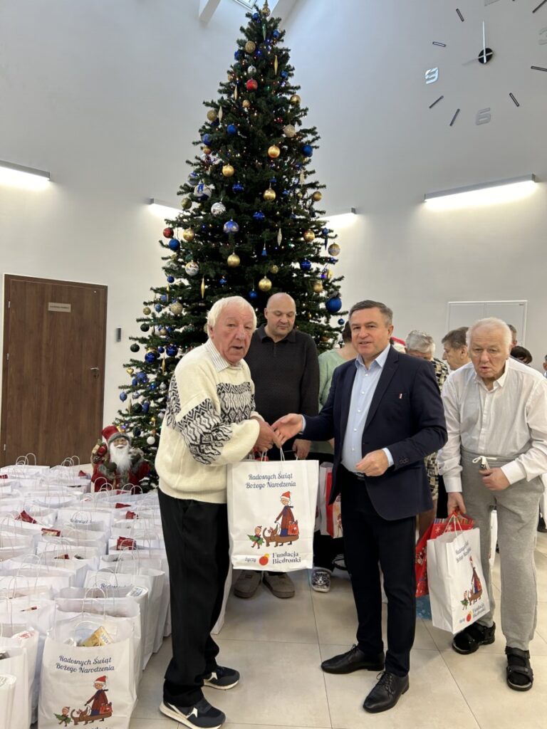 Uczestnicy wydarzenia wręczają prezenty z okazji świąt Bożego Narodzenia, stojąc przed dużą choinką w tle. Widać torby z darami i uśmiechnięte twarze, co podkreśla radosną atmosferę świąteczną.