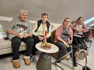 Grupa czterech kobiet w różnym wieku siedzi na kanapie w przytulnym pomieszczeniu. Na stoliku przed nimi znajduje się talerz z ciasteczkami i butelka napoju. Kobiety mają różne wyrazy twarzy, a jedna z nich trzyma kubek. W tle widoczna jest część wyposażenia pokoju.