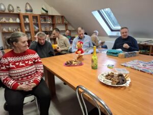 Spotkanie grupy osób przy stole w jadalni, z dekoracjami świątecznymi, kolorowymi ciastkami na talerzach oraz napojem w butelce. W tle widoczni uczestnicy w różnorodnych swetrach, cieszący się chwilą.