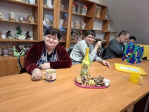 Grupa osób bawiących się przy stole w sali, na którym znajdują się ciastka, napój gazowany oraz kolorowe zabawki konstrukcyjne. W tle widoczne półki z dekoracjami i rękodziełem.