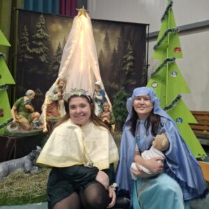 Dwie kobiety w strojach nawiązujących do postaci biblijnych pozują w pobliżu szopki bożonarodzeniowej z figurkami świętej rodziny. Tło z drzewami i ozdobami świątecznymi tworzy przytulną atmosferę.