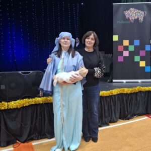 Kobieta przebrana za Maryję trzymająca dziecko oraz druga kobieta w czarnej bluzie, stojące na tle sceny z dekoracjami świetlnymi i banerem Podlaskie.