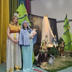 Kobiety w kostiumach anioła i Maryi stoją obok szopki bożonarodzeniowej, z figurkami przedstawiającymi scenę narodzenia Jezusa, w tle choinki i dekoracje świąteczne.