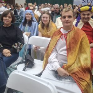 Grupa osób siedzących na widowni podczas wydarzenia, w tym kobieta w czarnej bluzie z kolorowymi haftami, mężczyzna w złotej pelerynie oraz kobieta w niebieskiej sukni. W tle widać tłum widzów.