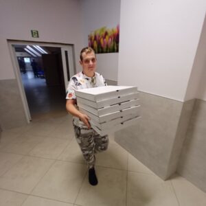 Mężczyzna niosący kilka pudełek pizzy w korytarzu, w tle widoczne kwiaty na ścianie.