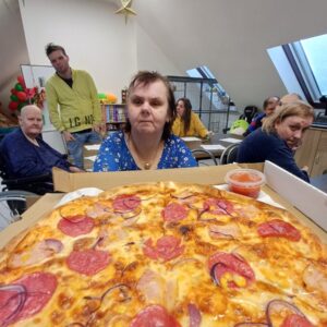 Grupa osób siedzących w sali z pizzą na pierwszym planie. Pizza z serem, pepperoni, cebulą i kukurydzą, obok sos pomidorowy. W tle widoczna dekoracja i inni uczestnicy spotkania.