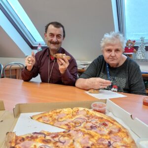 Mężczyzna i kobieta siedzą przy stole, uśmiechając się i trzymając kawałki pizzy. Na stole znajduje się duża pizza z szynką i cebulą oraz sosy. W tle widać okno oraz dekoracje świąteczne.